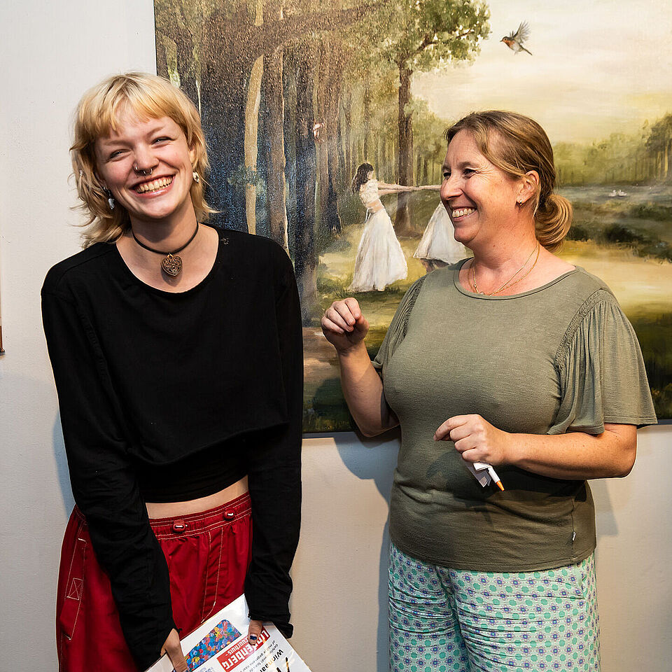 een jonge vrouw lacht met een certificaat in haar hand, naast haar staat een docent. Ze staan voor een kunstwerk.