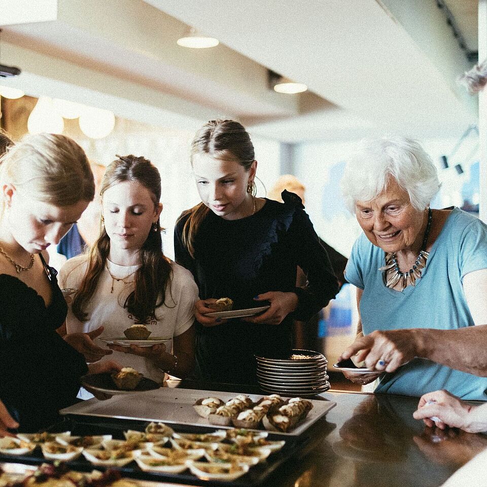 3 jonge meiden en een oudere dame staan rondom schalen met eten