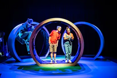 Drie acteurs staan in en rond grote, ronde decorstukken; de scène is kleurrijk verlicht in blauw en groen met speelse houding.