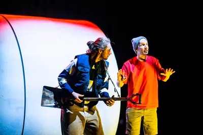 Twee acteurs staan op het podium: één met een schep als gitaar, de ander in rood shirt gebarend onder fel licht.