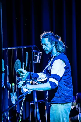 Een man met lang haar bespeelt een installatie van alledaagse voorwerpen zoals lepels en emmers, verlicht door blauw podiumlicht.
