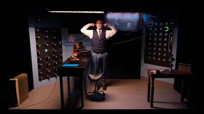 Een man in pak staat in een retro kantoor vol knoppen en kabels, met zijn vingers in zijn oren tegen het geluid.