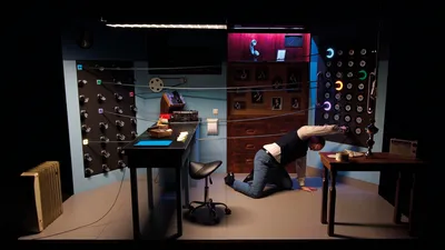 Een man knielt op de grond in het kantoor, terwijl hij aan een verlichte knop draait tussen talloze snoeren en lampjes.