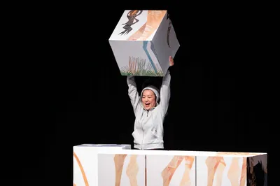 Een acteur tilt blij een groot beschilderd blok boven haar hoofd, terwijl andere blokken om haar heen opgestapeld staan.