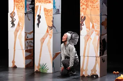 Een acteur in een grijs kostuum met olifantenoren kijkt lachend omhoog tussen torens van blokken met giraffenpoten.