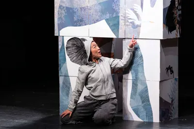 Een acteur knielt en wijst enthousiast omhoog naar een afbeelding op een toren van beschilderde blokken.