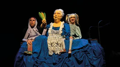 Een grote pop van een oudere vrouw in blauwe jurk zit tussen twee acteurs; ze houdt trots een maiskolf omhoog.