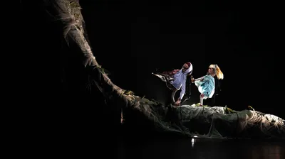 Twee acteurs balanceren op een kronkelende boomstam in een donker bosdecor, met dramatisch licht en nevelige doeken om hen heen.