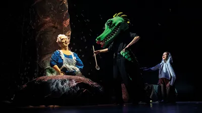 Een acteur met een groot krokodillenmasker staat met een houten lepel voor een oudere vrouw in blauwe jurk en een meisje.