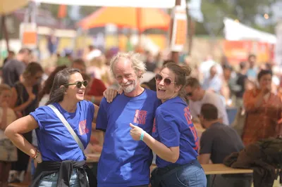 Drie vrijwilligers in blauwe shirts lachen samen op een druk festivalterrein.
