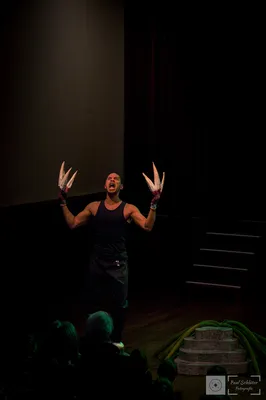 Een acteur met grote klauwachtige handen schreeuwt expressief op het podium tijdens een theatervoorstelling, terwijl het publiek toekijkt.