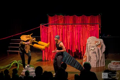 Twee spelers staan op een kleurrijk podium met rode doeken; één draagt een dinosaurusmasker, de ander heeft een staartkostuum.