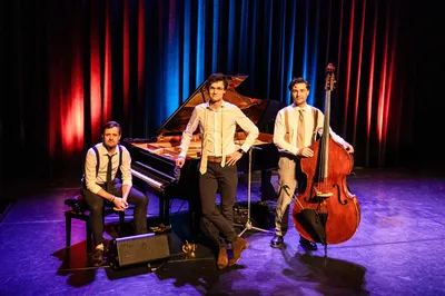 Drie muzikanten poseren op het podium voor piano en contrabas, in nette kleding en warme theaterverlichting met rood en blauw licht.