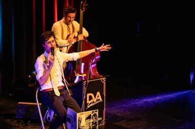 Een zanger zit op een kruk met microfoon in de hand, zingend terwijl de contrabassist achter hem meespeelt op een sfeervol verlicht podium.