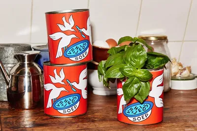 Twee blikken Vrijheidssoep zijn op elkaar gestapeld op het aanrecht. Een derde blik staat ernaast, net als een plantje basilicum en een schaal eieren.