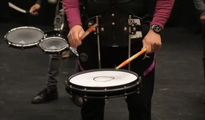 Een manslaat met drumstokken op een drum. Op de achtergrond staan nog meer drummers.
