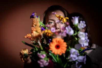 Een vrouw kijkt ernstig recht in de camera, half verscholen achter een grote bos bloemen met oranje, paarse en gele tinten.