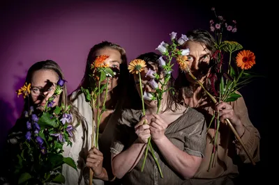 Vier vrouwen staan dicht bij elkaar en houden bloemen voor hun gezichten, waardoor hun ogen deels zichtbaar zijn in gekleurd licht.