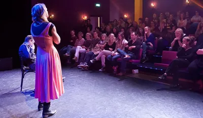 Publiek kijkt in de Karolingenzaal naar twee mensen die optreden op het podium