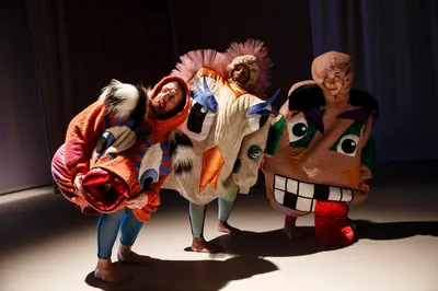 Dansers in kleurrijke, speelse kostuums met fantasiegezichten buigen naar voren op een zacht verlichte toneelvloer