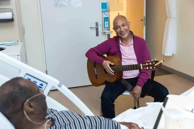 Een muzikant met zijn gitaar op schoot lacht naar een patiënt in een ziekenhuisbed.