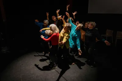 Een groep oudere dansers in kleurrijke kleding staat bij elkaar in het midden van het podium. Ze bewegen verschillende kanten op.