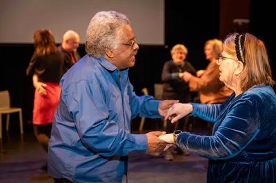 Een oudere man en vrouw dansen samen. Ze hebben elkaars handen vast en lachen. Op de achtergrond dansen meer paren.