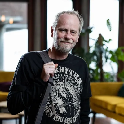 Man met schoudertas kijkt vriendelijk in de camera in een lichte loungeruimte.