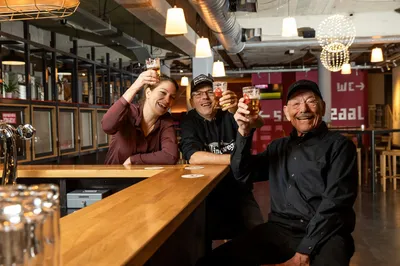 Drie mensen proosten vrolijk met een glas bier aan de bar.
