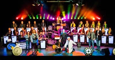 Het Nijmeegs Amusementsorkest speelt op een kleurrijk verlicht podium met zangers vooraan, instrumenten achter hen en grote klokken als decor.