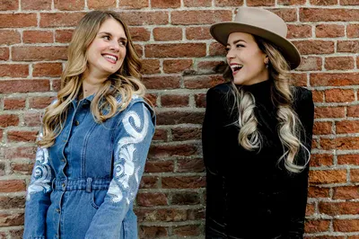 Twee vrouwen staan lachend voor een bakstenen muur.