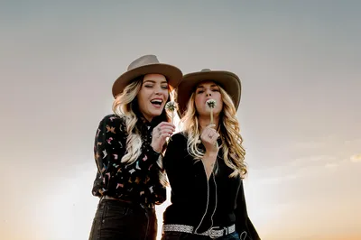 Twee vrouwen met een cowboyhoed op blazen de pluisjes van een paardenbloemsteel.