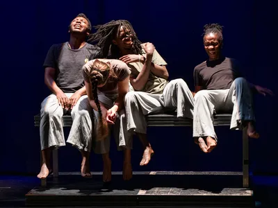 Vier dansers zitten op een bank en kijken intens in helder podiumlicht.