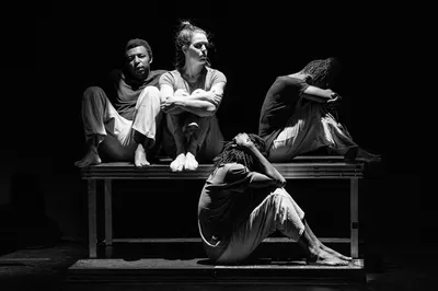 Vier dansers zitten in stilte op een verhoogd platform, dramatisch verlicht.