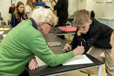 Een oudere dame in een groene trui leunt over een tafel en tekent iets met potlood op papier. Een jonge jongen met zwarte trui kijkt mee naar wat zij tekent.