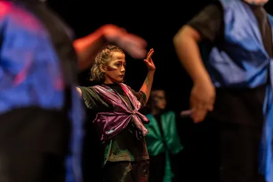 Een kind op een podium danst geconcentreerd, gekleed in een paars kostuum met felgekleurde schmink op het gezicht