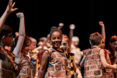 Groep kinderen in baksteenpatronen kostuums viert een optreden, met centraal een lachend meisje met zwarte strepen op haar wangen