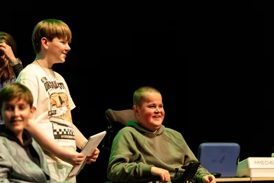 Jongeren op een podium lachen en presenteren, met één deelnemer in een rolstoel op de voorgrond