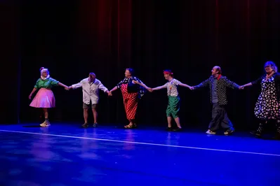 Op een podium loopt een rij mensen in bontgekleurde kleding. Ze hebben elkaars handen vast.