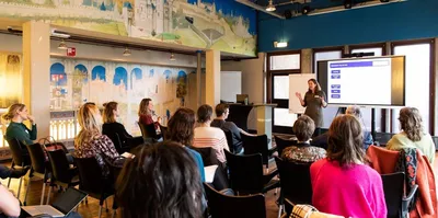 In de valkhofzaal zitten mensen in rijen op stoelen. Een vrouw geeft een presentatie met achter haar en groot beeldscherm.