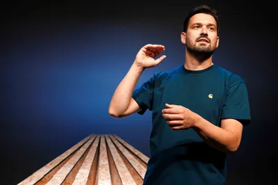 Jasper van der Veen staat op een houten podium en maakt een sprekend gebaar met zijn hand, tegen een donkerblauwe achtergrond.