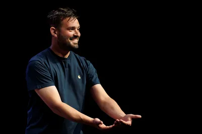 Jasper van der Veen glimlacht en reikt met open handen naar voren op een donker podium.