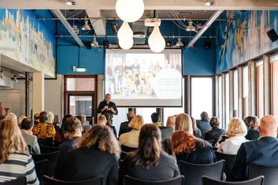 In de Valkhofzaal zitten mensen in rijen op stoelen. Een man geeft een presentatie met achter hem en groot beeldscherm.