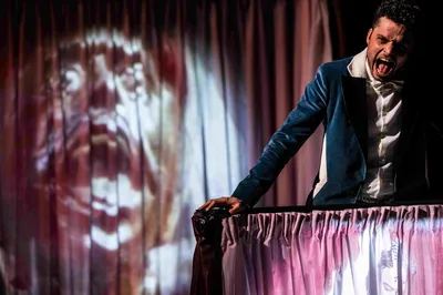 Een acteur in een blauw colbert schreeuwt expressief boven een theaterdoek, terwijl een groot, angstig gezicht op de achtergrond wordt geprojecteerd.