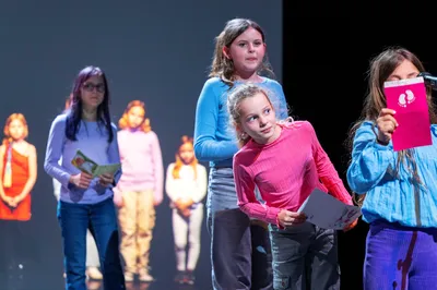 Vier kinderen staan op een podium en lezen voor uit kaarten; op de achtergrond zijn meer kinderen geprojecteerd op een scherm.