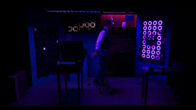 Het kantoor is donkerblauw verlicht; een man loopt langzaam rond terwijl felle paarse ringen op een wand vol lampjes gloeien.