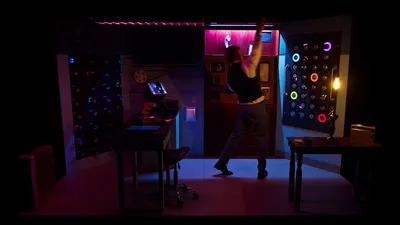 Een man strekt zijn armen omhoog in een kamer vol gekleurde lichten en draait aan een muur vol ronde knoppen.