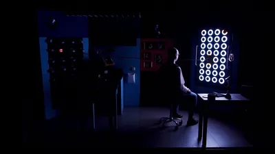 Een man zit stil op een kruk, omgeven door duisternis, en kijkt naar tientallen helderwitte ringen die oplichten aan de muur.