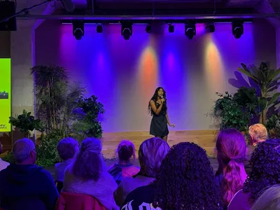 Een zangeres staat onder gekleurde lichten te zingen. Op de voorgrond zitten mensen te kijken. 