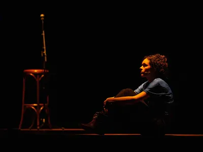 Nora zit met haar handen over haar knieën op het podium en kijkt in de verte. 